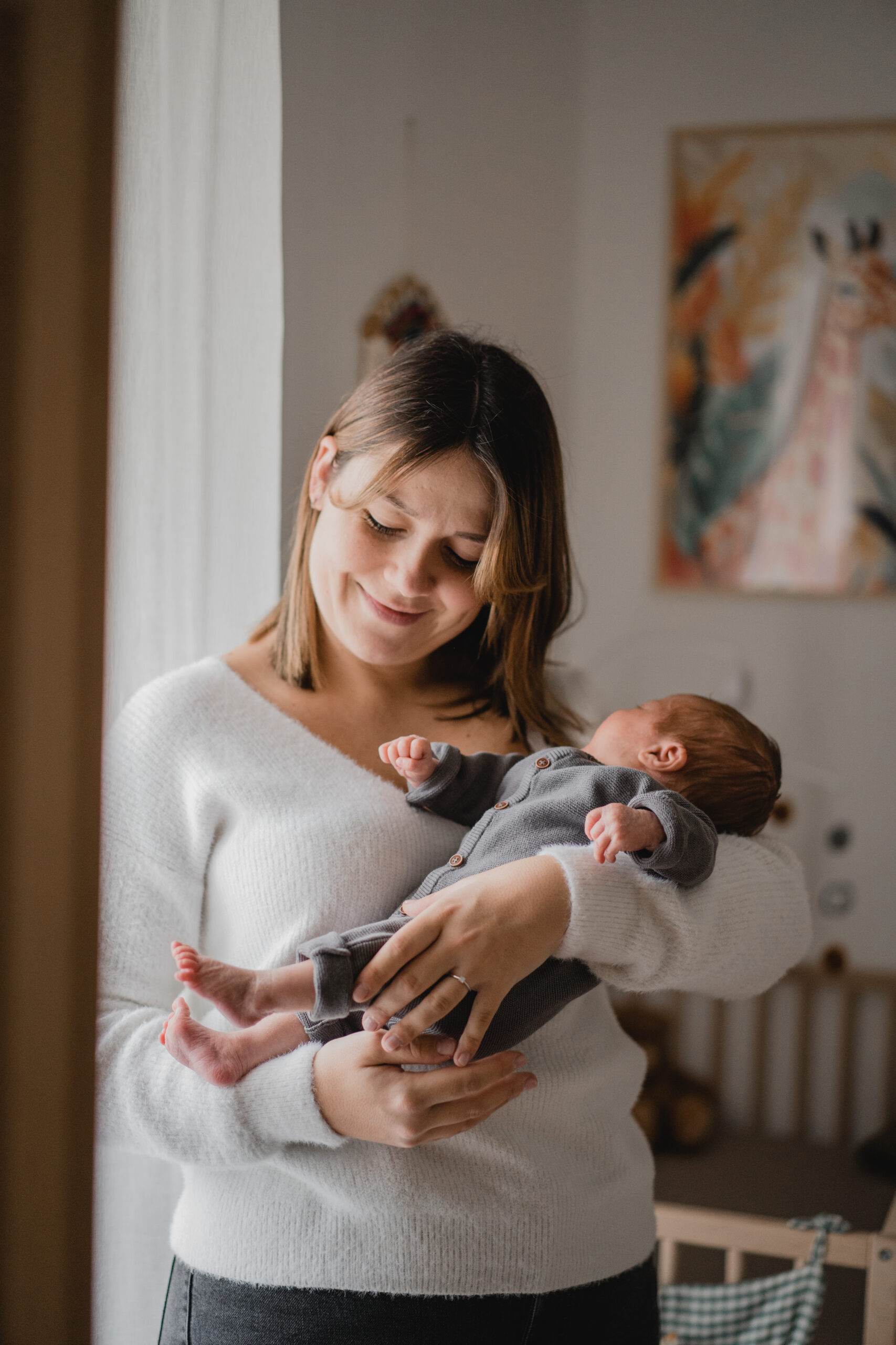 Jeune maman berçant son nouveau-né dans sa chambre pendant une séance photo naissance.