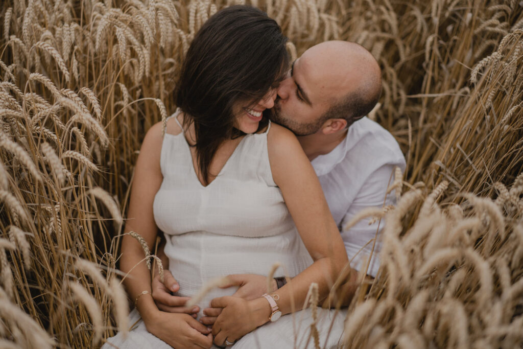 Moment de tendresse dans un champ de blé où un futur papa embrasse la future maman enceinte.