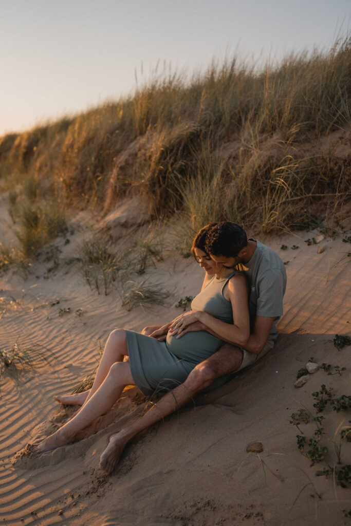 Séance grossesse au coucher du soleil sur les dunes, future maman assise contre son compagnon.
