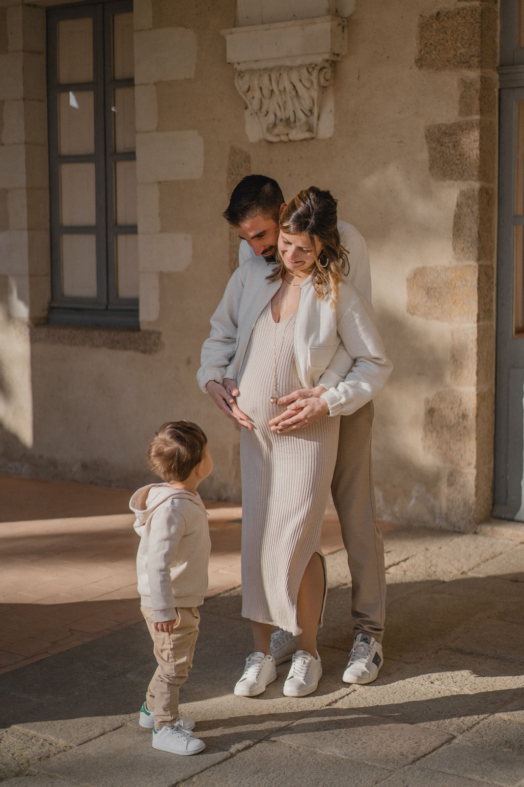 Jeune famille réunie autour du ventre rond de la future maman devant une bâtisse en pierre.