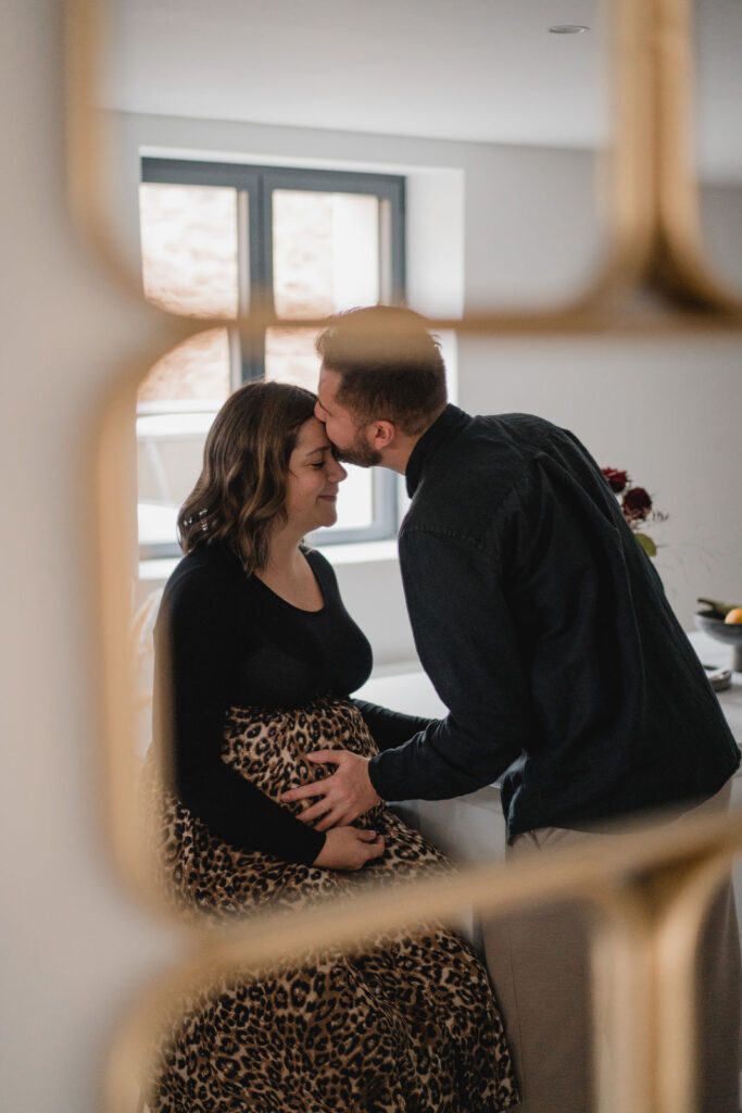 Futur papa embrassant la future maman enceinte à travers le reflet d'un miroir lors d'une séance à domicile.