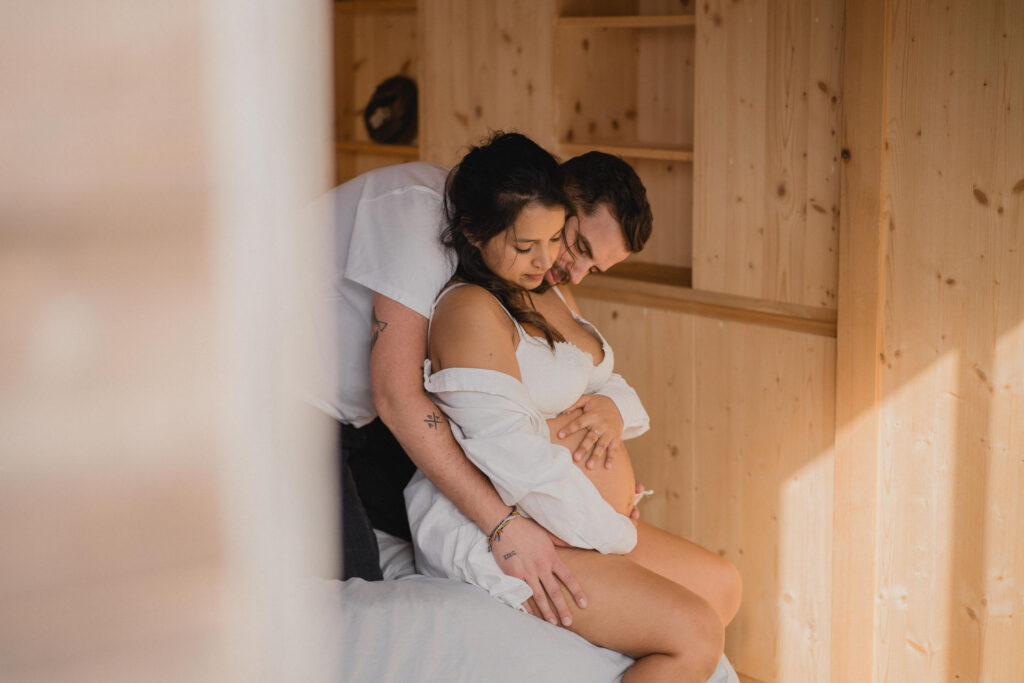 Futur papa enlacé à la future maman enceinte dans une chambre baignée de lumière naturelle.