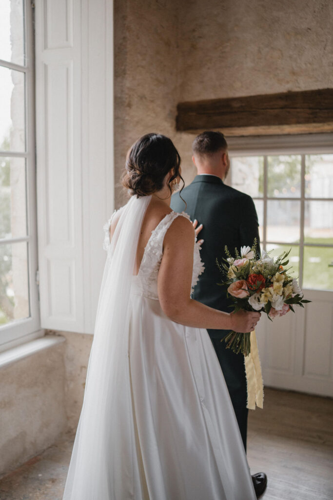 La mariée s'apprête à tapoter l'épaule de son futur mari, placé de dos, pour qu'il se retourne et la découvre dans sa robe blanche.