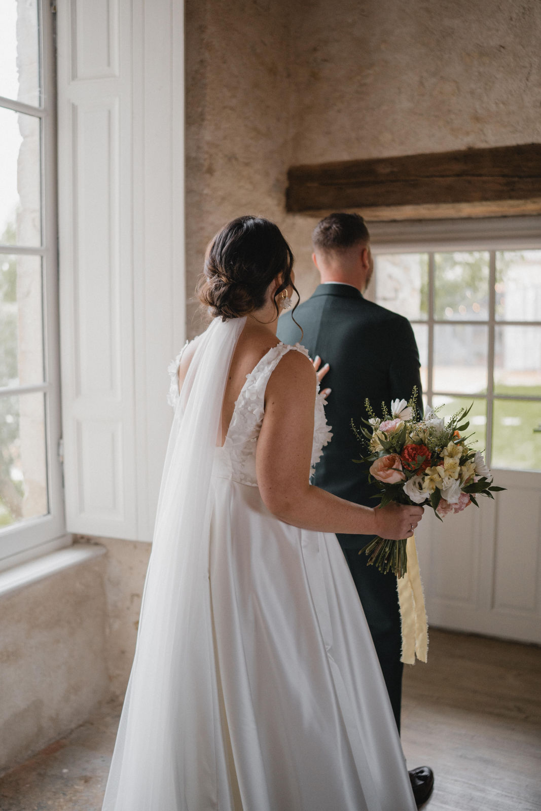 La mariée s'apprête à tapoter l'épaule de son futur mari, placé de dos, pour qu'il se retourne et la découvre dans sa robe blanche.