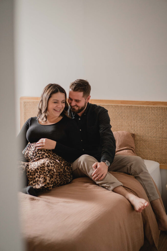 Futurs parents installés sur un lit lors d'une séance photo grossesse à domicile.